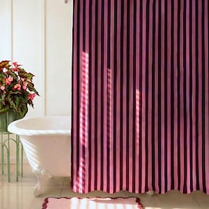 Puede incluir: Un cuarto de baño con una cortina de ducha a rayas rosas y burdeos. Una bañera blanca con patas de león y una alfombra de baño rosa con volantes están frente a la cortina. Una planta en maceta con flores rosas está cerca de una ventana.