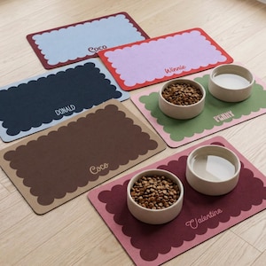 May include: A collection of rectangular pet placemats with scalloped edges in various colors including light blue, lavender, dark blue, olive green, brown, and maroon. Each mat features a personalized name: Coco, Winnie, Donald, Penny, and Valentine.