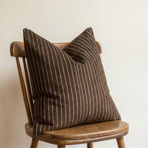 May include: A square throw pillow featuring a dark brown fabric with a vertical pinstripe pattern in a lighter tan color. The pillow has a soft, textured appearance and is positioned upright, showcasing its full shape and consistent striped design.
