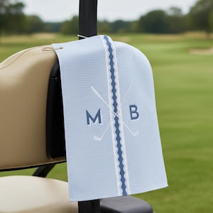 May include: A light blue waffle-textured golf towel featuring a vertical navy blue wavy stripe down the center. The design includes two crossed white golf club icons and the navy blue capital letters M and B positioned on either side.