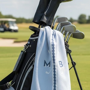 May include: A light blue golf towel with a waffle texture, featuring the embroidered letters M and B alongside a crossed golf club graphic. The towel hangs from a black golf bag containing several golf clubs with black protective head covers.