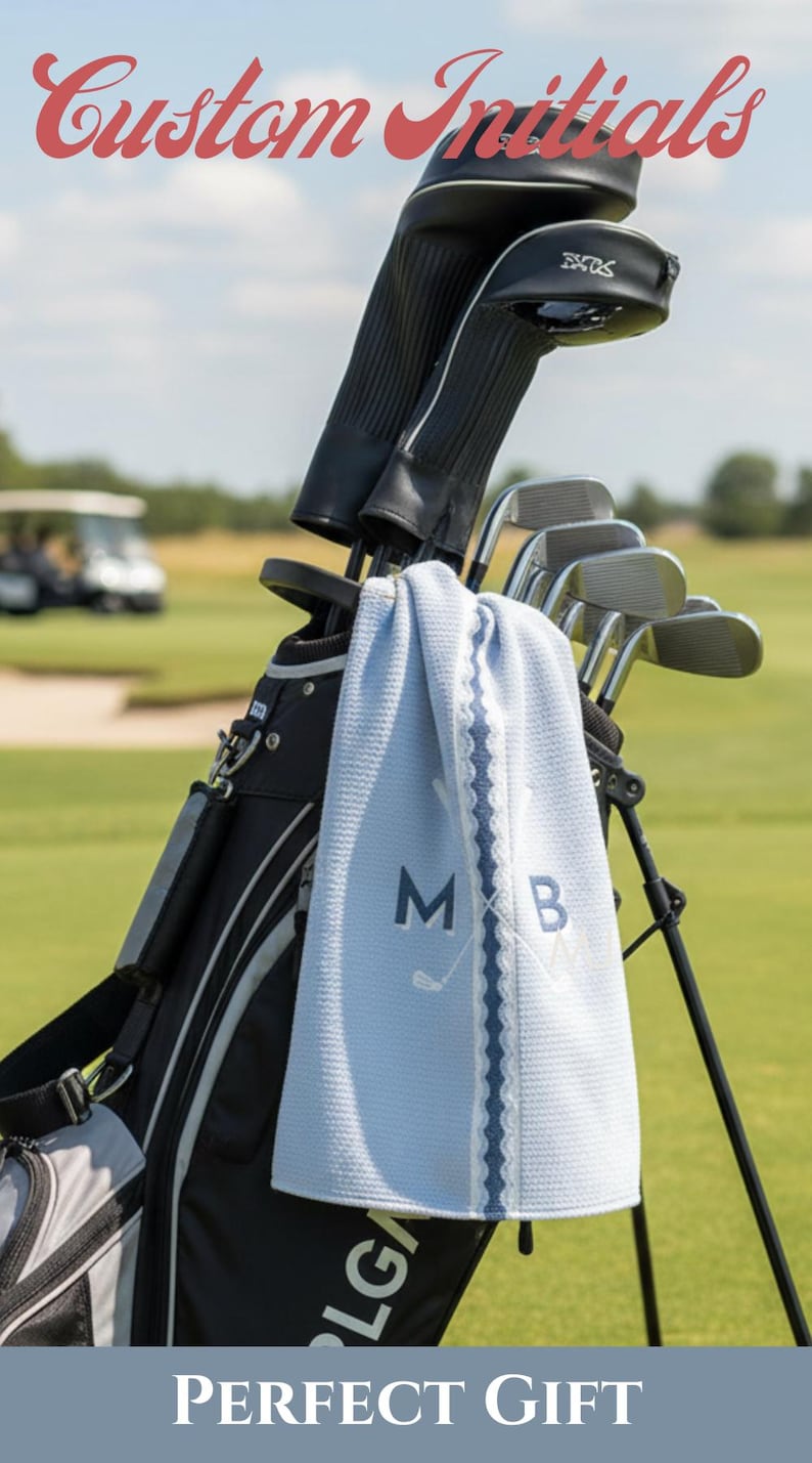May include: A light blue golf towel with a waffle texture, featuring the embroidered letters M and B alongside a crossed golf club graphic. The towel hangs from a black golf bag containing several golf clubs with black protective head covers.