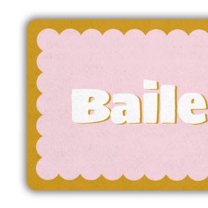 May include: A rectangular card with a scalloped edge. The card has a light pink center with the word "Baile" in bold white letters. The border is a golden yellow color.