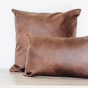 May include: Two brown faux leather throw cushions. One is square, the other is rectangular. The cushions have a textured surface and are placed on a wooden surface against a white background. The cushions are a warm, earthy tone.