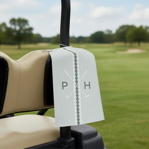 Custom golf towel with monogram initials and crossed golf club graphic, hanging on a golf cart seat. This personalized golf accessory is perfect for cleaning clubs and makes a unique golf gift for men, fathers, husbands, and golf enthusiasts.