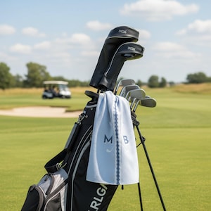 May include: A black golf bag containing several golf clubs with black headcovers. A light blue textured golf towel hangs from the bag, featuring a dark blue vertical stripe, the letters M and B, and a graphic of two crossed golf clubs in white.