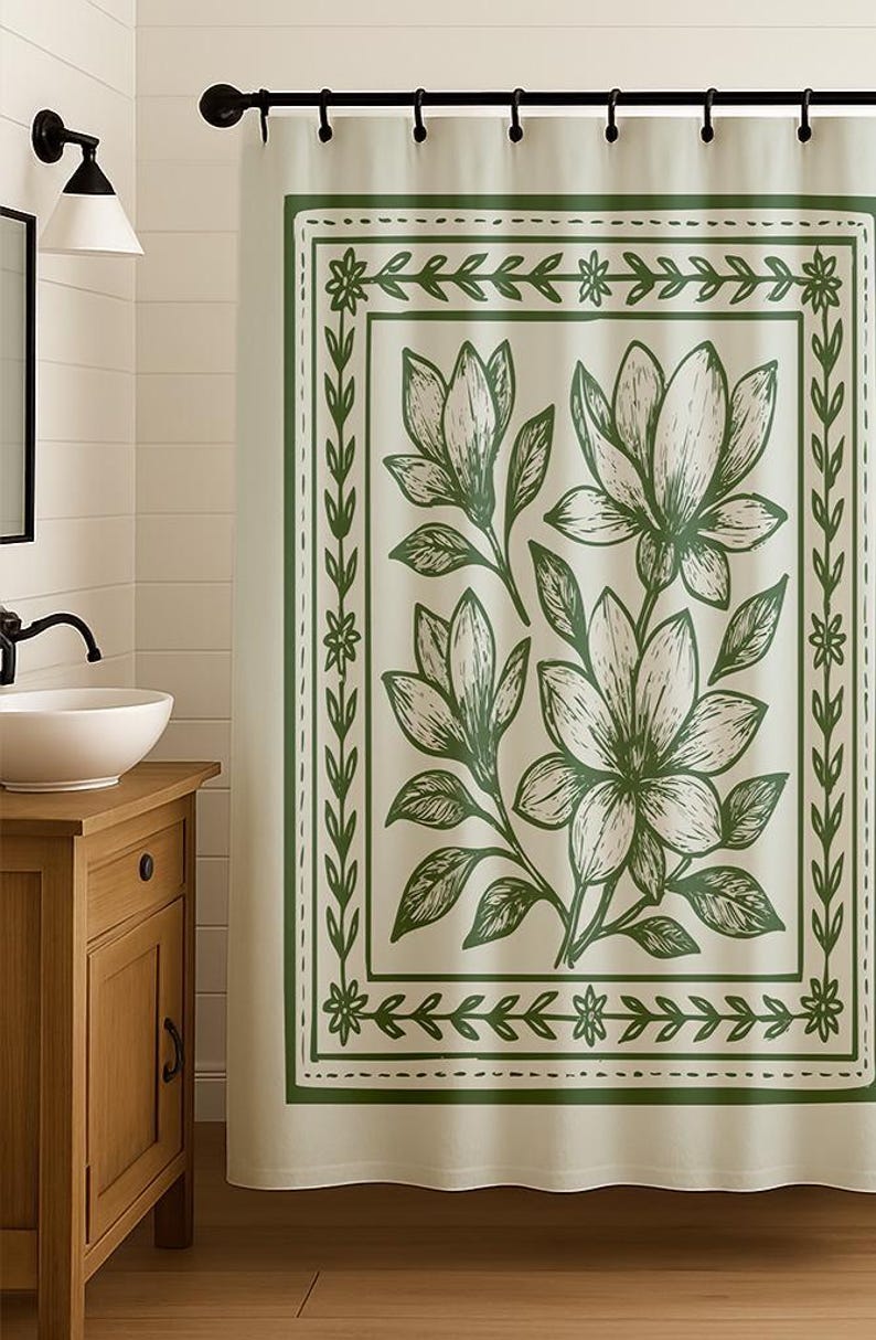 May include: A cream-colored shower curtain with a green floral design. The design features large magnolia flowers and leaves within a green border. The curtain is hanging in a bathroom setting, with a wooden vanity and a white sink visible.