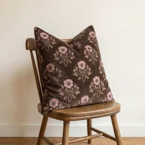 May include: A square throw cushion featuring a dark brown velvet fabric with a repeating pattern of light pink flowers and green leaves. The cushion is plump and has a soft texture, showcasing a floral botanical print across the entire surface.