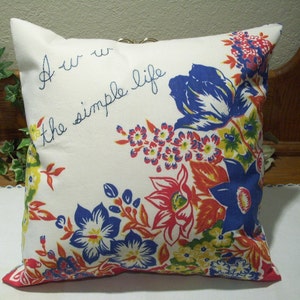 May include: Square pillow with a white background and floral pattern in red, blue, yellow, and orange. The pillow has the embroidered text "Aw the simple life". The pillow has a red trim.