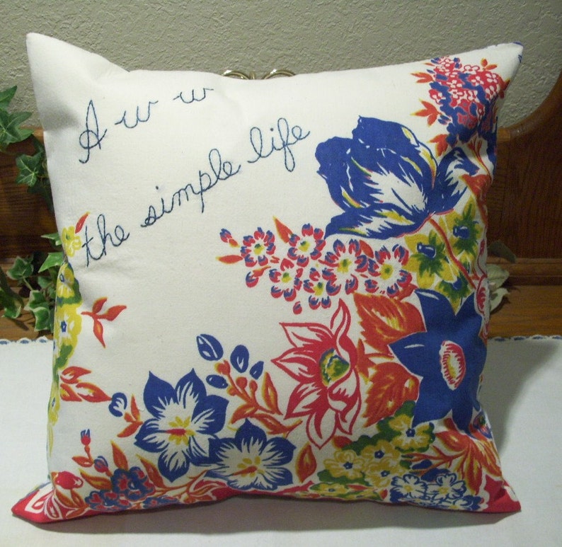 May include: Square pillow with a white background and floral pattern in red, blue, yellow, and orange. The pillow has the embroidered text "Aw the simple life". The pillow has a red trim.