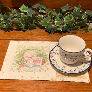 May include: A white teacup and saucer with blue floral and vine details sit on a placemat with a garden scene. The placemat features a white wicker chair and the text "Screen Door Views". The cup and saucer have a blue trim.