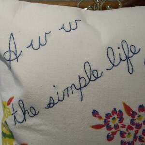 May include: A white pillow with blue embroidered text that reads "Aw the simple life." The pillow features a floral pattern with blue, red, and yellow flowers. The pillow is a decorative item.