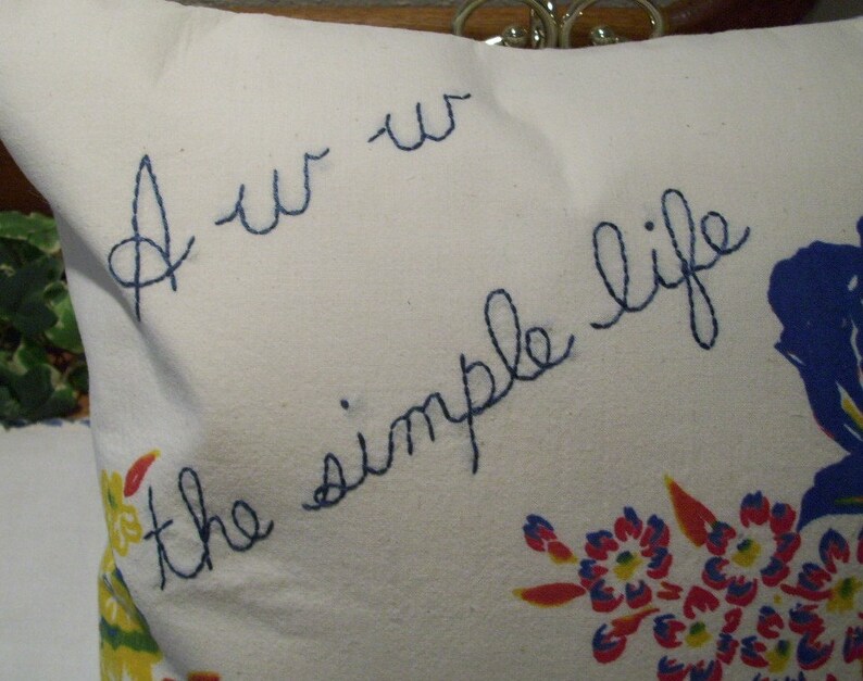 May include: A white pillow with blue embroidered text that reads "Aw the simple life." The pillow features a floral pattern with blue, red, and yellow flowers. The pillow is a decorative item.