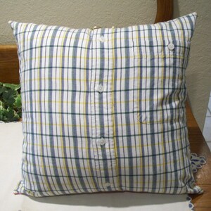 May include: Square pillow with a plaid pattern in white, blue, yellow, and green. The pillow cover is made from a button-down shirt, with the buttons and pocket visible. The pillow is on a wooden bench.