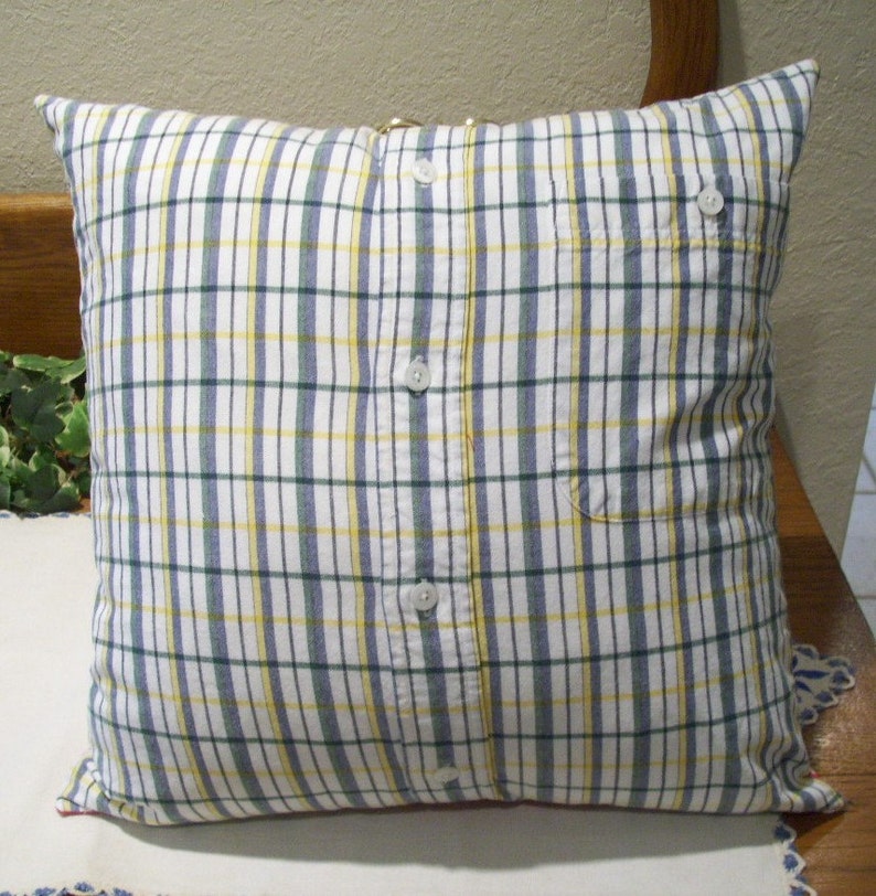 May include: Square pillow with a plaid pattern in white, blue, yellow, and green. The pillow cover is made from a button-down shirt, with the buttons and pocket visible. The pillow is on a wooden bench.