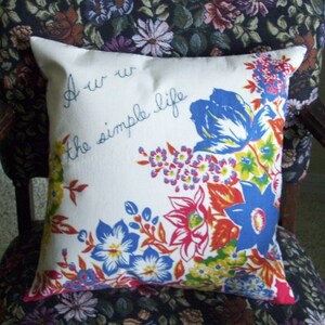 May include: A white square pillow with a floral design in blue, red, yellow, and orange. The pillow has the text "Aw w the simple life" in blue script. The pillow is sitting on a floral patterned chair.