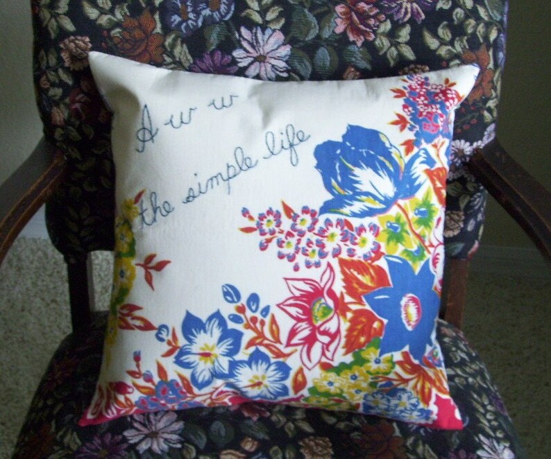 May include: A white square pillow with a floral design in blue, red, yellow, and orange. The pillow has the text "Aw w the simple life" in blue script. The pillow is sitting on a floral patterned chair.