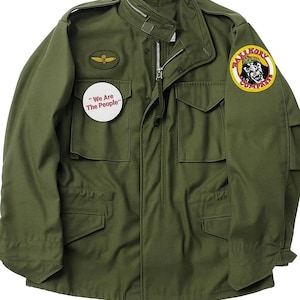 May include: Olive green military-style jacket with a zip-up front and multiple pockets. Features a round patch with a graphic and the text "Rattikory Company". A small patch with a gold emblem and a white circular patch with the text "We Are The People" are also visible.