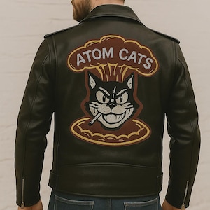 May include: Black leather jacket with a "Atom Cats" patch on the back. The patch features a cartoon cat smoking a cigarette, with the words "ATOM CATS" above. The jacket has a classic biker style with a zipper closure and a notched collar.