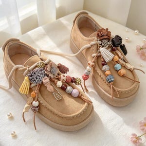 May include: A pair of tan suede shoes featuring thick laces adorned with various decorative charms, including small fabric flowers, colorful wooden and plastic beads, tassels, and a metal safety pin attached to the front of each shoe.