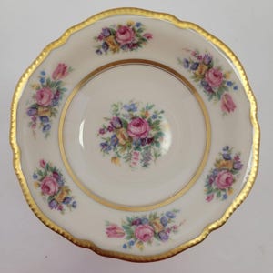 Può includere: Un piatto di porcellana bianca con bordo dorato smerlato e fascia interna dorata. Il piatto è decorato con grappoli di rose rosa, fiori viola, gialli e blu. Il motivo floreale si ripete sul bordo e al centro.