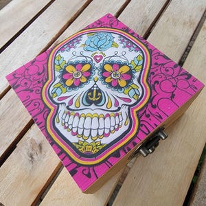 May include: A square wooden box featuring a vibrant sugar skull design on the lid. The skull is decorated with bright yellow, pink, and light blue floral patterns against a white background, surrounded by a bold pink border with black scrollwork.