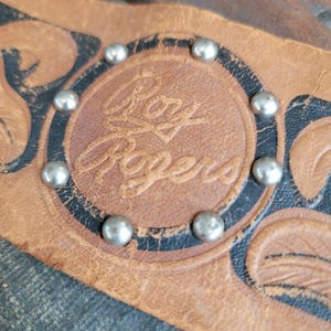 Op de afbeelding: Close-up van een versleten leren item met de in reliëf aangebrachte tekst "Roy Rogers" in een cirkelvormig ontwerp. Het ontwerp is omgeven door zilverkleurige studs. Het leer is bruin met zwarte accenten.