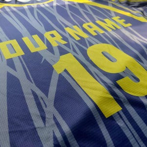 May include: A blue athletic jersey featuring a yellow graphic pattern of thin, intersecting lines. The shirt displays the text YOUR NAME in yellow block letters above the large yellow number 19, printed on a textured, breathable fabric surface.