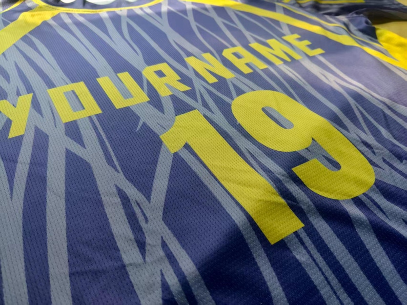May include: A blue athletic jersey featuring a yellow graphic pattern of thin, intersecting lines. The shirt displays the text YOUR NAME in yellow block letters above the large yellow number 19, printed on a textured, breathable fabric surface.