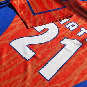 May include: A sports jersey featuring a vibrant orange and blue color scheme with a subtle abstract pattern. The back of the garment displays the large white number 21 and the partial text NATA printed in a bold, clean font across the upper section.