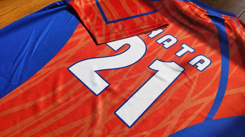 May include: A sports jersey featuring a vibrant orange and blue color scheme with a subtle abstract pattern. The back of the garment displays the large white number 21 and the partial text NATA printed in a bold, clean font across the upper section.