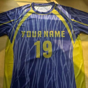 May include: A short-sleeve athletic jersey in purple and yellow featuring a light purple abstract line pattern. The front displays the text YOUR NAME in yellow block letters above the large yellow number 19 centered on the chest area.