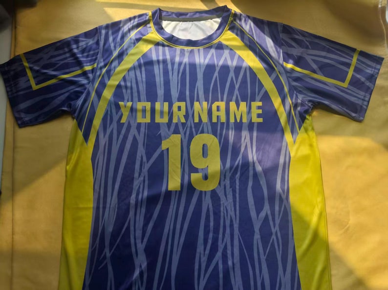 May include: A short-sleeve athletic jersey in purple and yellow featuring a light purple abstract line pattern. The front displays the text YOUR NAME in yellow block letters above the large yellow number 19 centered on the chest area.