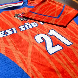 May include: A bright orange and blue athletic jersey featuring a textured pattern, the text SESI S&Atilde;O PAULO, and the number 21 in white with a blue outline. The shirt has a crew neckline and short sleeves with blue accents on the shoulders and sides.