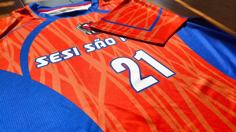 May include: A bright orange and blue athletic jersey featuring a textured pattern, the text SESI S&Atilde;O PAULO, and the number 21 in white with a blue outline. The shirt has a crew neckline and short sleeves with blue accents on the shoulders and sides.