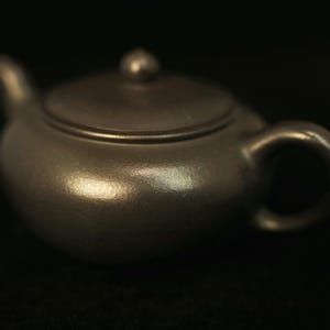 May include: A small, brown ceramic teapot with a rounded body, a curved handle, and a small spout. The lid has a small knob on top. The teapot is set against a dark background, highlighting its smooth, reflective surface.