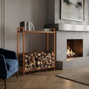 May include: A rectangular metal firewood rack with a copper-toned finish, featuring a flat top shelf holding several books and a lower section filled with stacked, split logs. The structure is open-sided with thin vertical and horizontal bars.