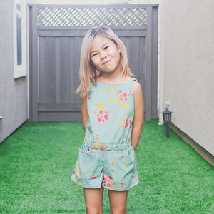 Puede incluir: Una ni&ntilde;a con cabello rubio lleva un mono azul claro con un dise&ntilde;o de pintura salpicada de colores. Est&aacute; de pie en un patio trasero con hierba verde.