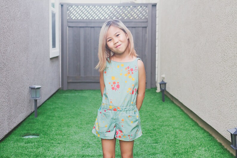 Puede incluir: Una ni&ntilde;a con cabello rubio lleva un mono azul claro con un dise&ntilde;o de pintura salpicada de colores. Est&aacute; de pie en un patio trasero con hierba verde.