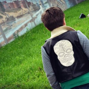 May include: A young person wearing a black leather vest with a white skull design on the back. The vest has a gray quilted lining and is worn over a green t-shirt.