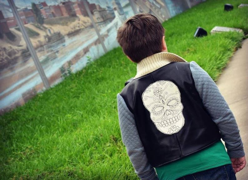 May include: A young person wearing a black leather vest with a white skull design on the back. The vest has a gray quilted lining and is worn over a green t-shirt.