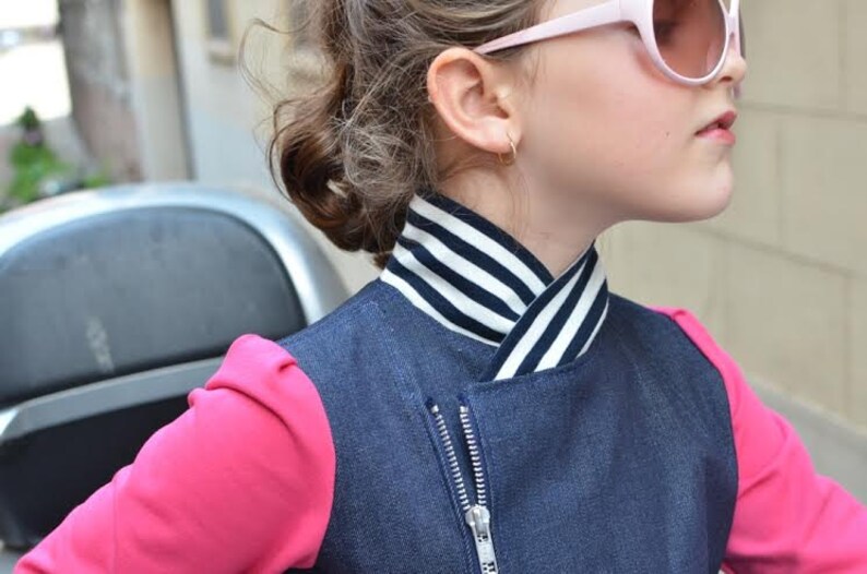 May include: A young person wearing a blue denim vest with a black and white striped collar, a pink long-sleeved shirt, and pink sunglasses.