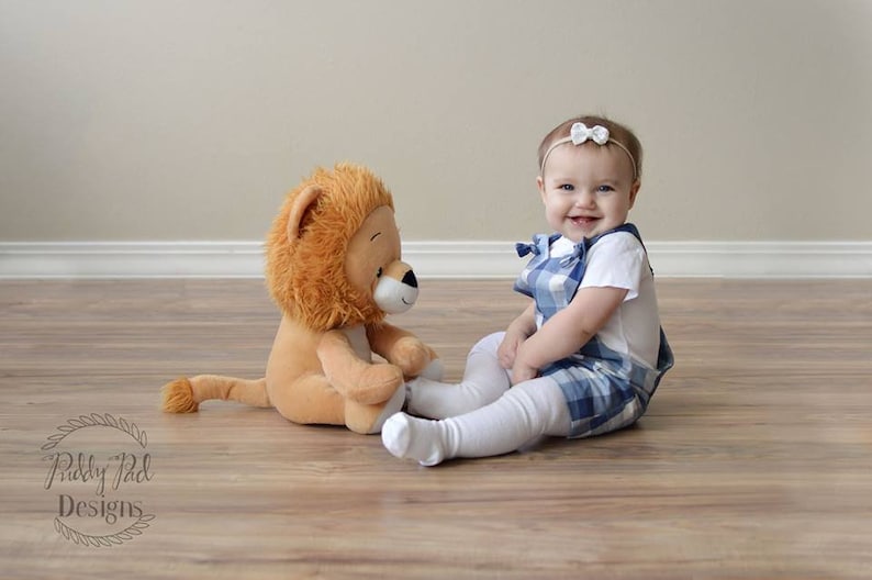 K&ouml;nnte beinhalten: Ein Baby in einem blauen und wei&szlig;en karierten Latzkleid und einem wei&szlig;en Stirnband mit Schleife sitzt auf einem Holzboden mit einem Pl&uuml;schl&ouml;wenspielzeug.