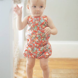 Puede incluir: Una ni&ntilde;a peque&ntilde;a con un mono de flores rojo y blanco y zapatos dorados. La ni&ntilde;a est&aacute; de pie sobre un suelo de madera.