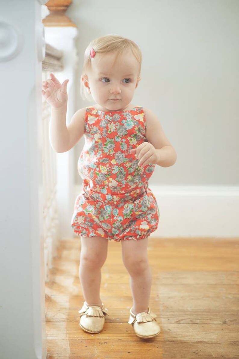 Puede incluir: Una ni&ntilde;a peque&ntilde;a con un mono de flores rojo y blanco y zapatos dorados. La ni&ntilde;a est&aacute; de pie sobre un suelo de madera.