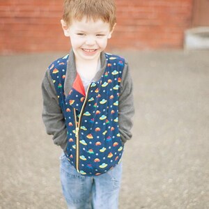 May include: A young boy wearing a blue and grey jacket with a colorful pattern of flying saucers. He is smiling and has his hands in his pockets. He is wearing blue jeans and green shoes.