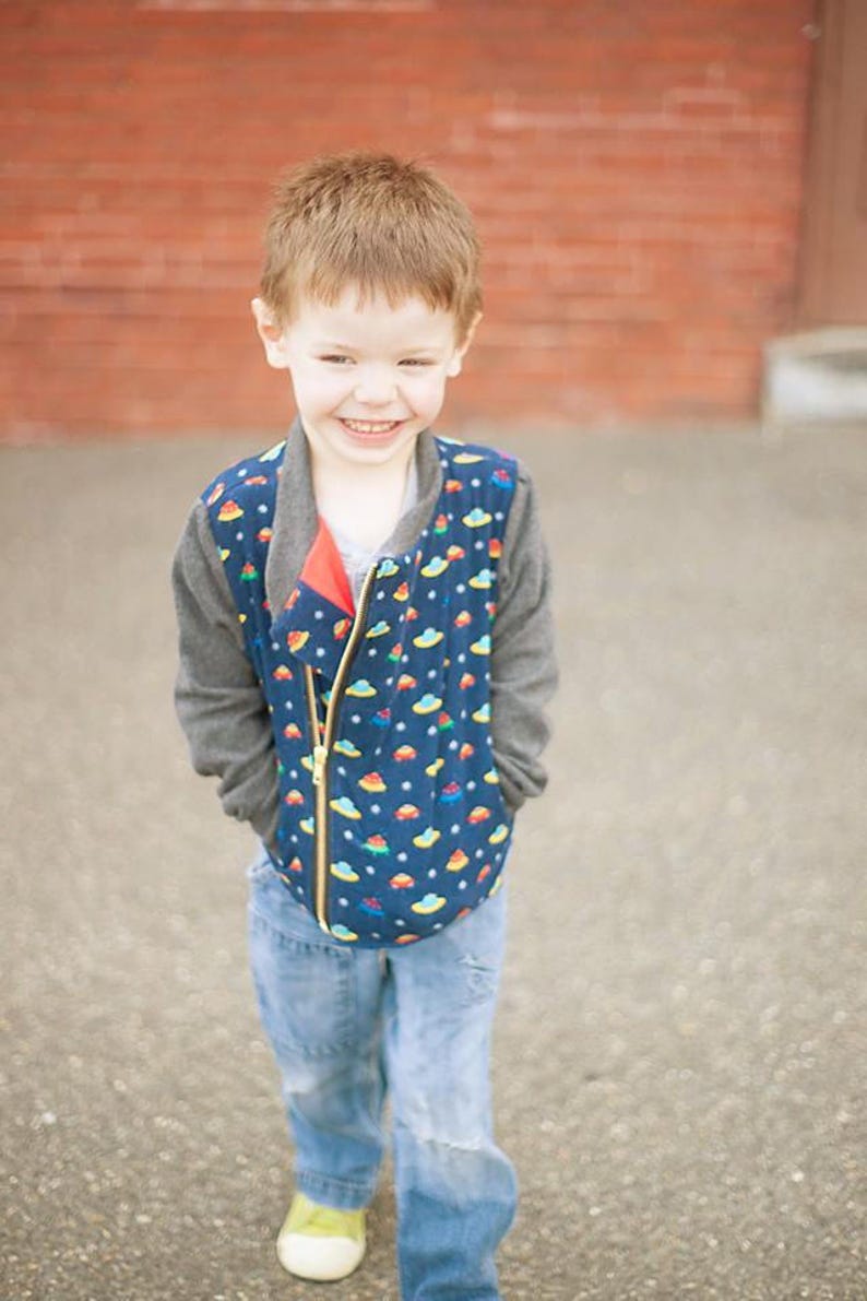 May include: A young boy wearing a blue and grey jacket with a colorful pattern of flying saucers. He is smiling and has his hands in his pockets. He is wearing blue jeans and green shoes.