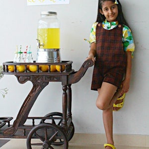 K&ouml;nnte beinhalten: Ein M&auml;dchen in einem karierten Kleid und einem Blumenhemd steht neben einem Holzkarren mit einem Krug Limonade und Zitronen. Ein Schild &uuml;ber dem Karren lautet "Lemonade 5&cent;".