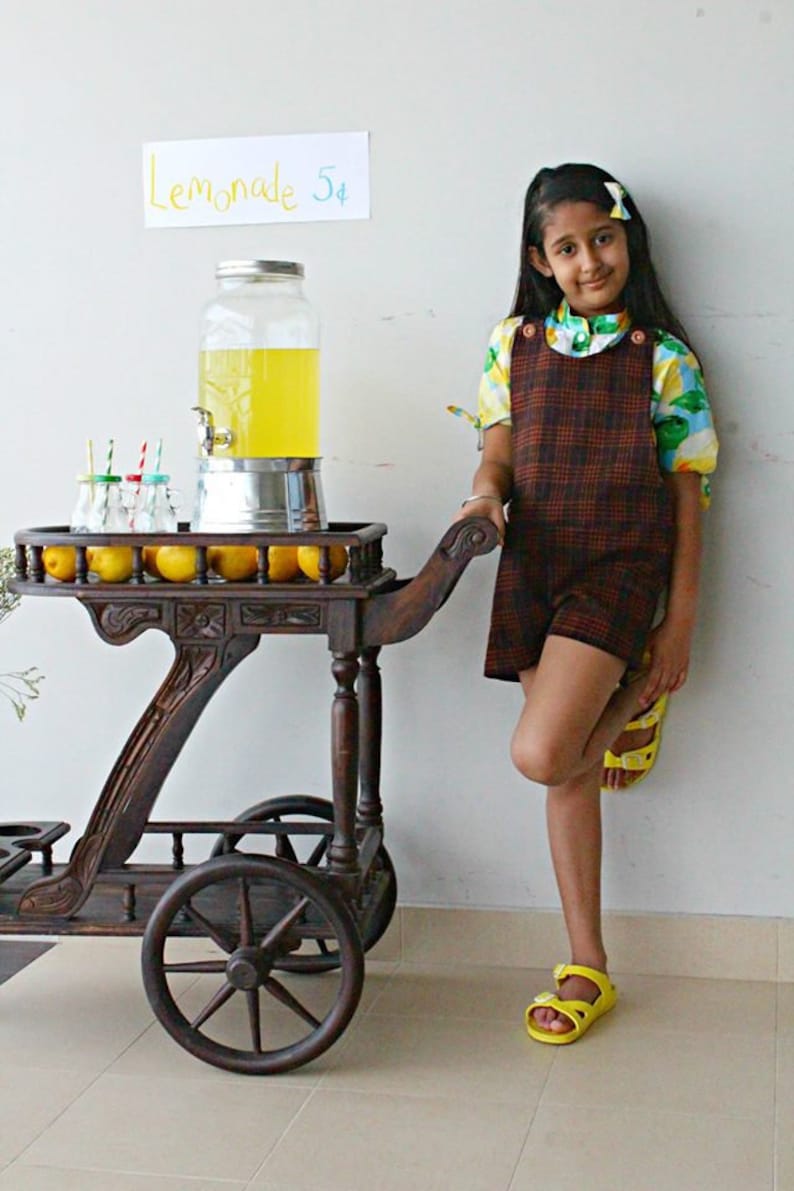 K&ouml;nnte beinhalten: Ein M&auml;dchen in einem karierten Kleid und einem Blumenhemd steht neben einem Holzkarren mit einem Krug Limonade und Zitronen. Ein Schild &uuml;ber dem Karren lautet "Lemonade 5&cent;".