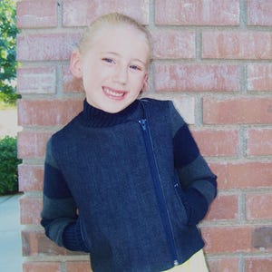 May include: A young person wearing a dark blue denim jacket with black and grey striped sleeves. The jacket has a zipper down the front and a collar.